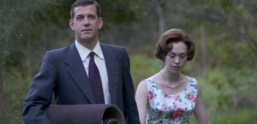 A man in a business suit carries a rolled up carpet and walks in the bush with a woman wearing a pearl necklace and 1960s-style dress