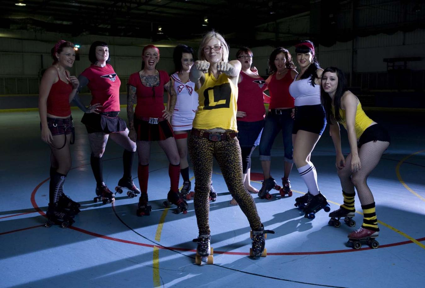 Nine female roller derby players wearing roller skates and standing in a semi-circle, posing. The middle player stands in front of the others and has her arms outstretched and hands bunched into fists.