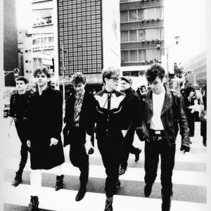 Publicity image of the six band members of INXS walking across a pedestrian crossing with tall buildings in the background.