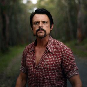 upper body shot of a menacing looking man standing in a road near a forest.