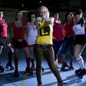 Nine female roller derby players wearing roller skates and standing in a semi-circle, posing. The middle player stands in front of the others and has her arms outstretched and hands bunched into fists.